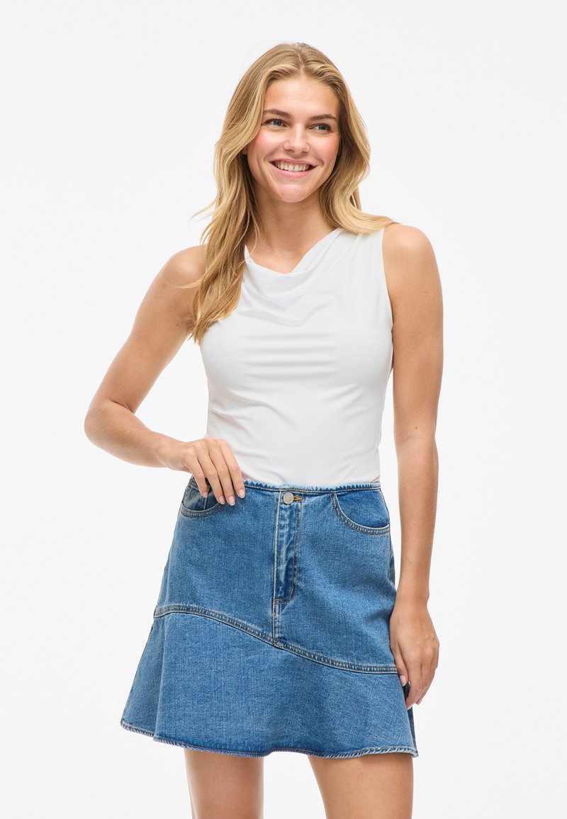 Smiling woman with long blonde hair wearing sleeveless white top and blue denim skirt, standing against white background.