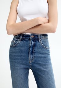 Jeans de mezclilla azul de tiro alto con lavado desgastado, cinco bolsillos y cierre de botón metálico, diseñados para una silueta ajustada.