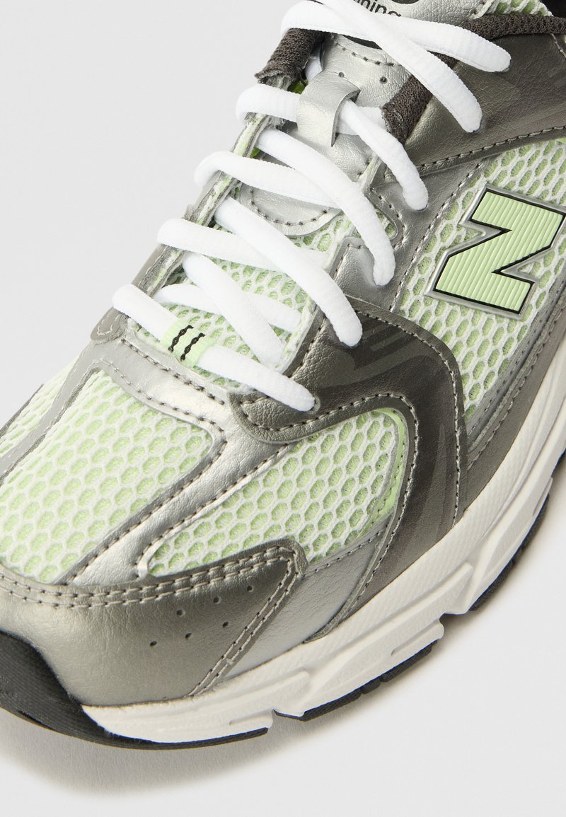 Chaussure de sport grise et vert clair avec des textures en maille et en cuir, présentant des lacets blancs et un logo "N" bien visible sur le côté.