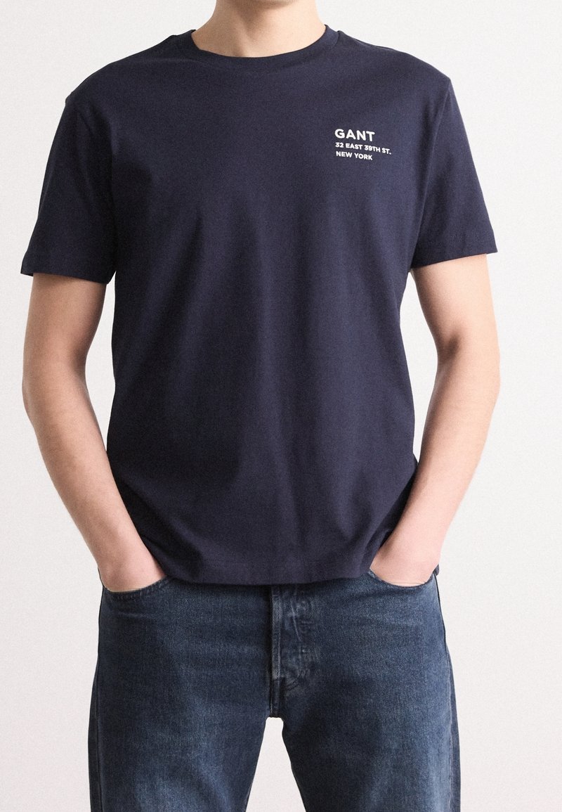 Homme portant un t-shirt GANT bleu marine avec un texte blanc "GANT 32 East 39th St. New York" sur le côté gauche de la poitrine, associé à un jean bleu foncé.