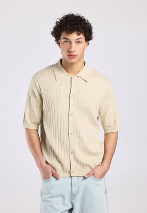 Jonge man met krullend haar, draagt een beige geribd kort mouw knopoverhemd en lichtblauwe spijkerbroek, handen in de zakken, neutrale achtergrond.