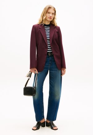 Naine, kes kannab maroonpunast jopet, triibulist särki, siniseid teksapükse, musti kontsakingi, hoides musta käekotti triibulise rihmaga, seisab valge tausta ees.