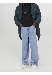 Grüne Pufferjacke mit Kapuze, schwarzes Reißverschluss-Sweatshirt und hellblaue Schlagjeans. Weiße Sneakers runden das Outfit ab.