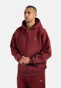 Sudadera con cremallera de color burdeos, hecha de tejido suave, que cuenta con una gran capucha, bolsillos tipo canguro, puños acanalados y una discreta etiqueta con logotipo en el pecho.