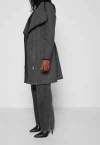 Persoon in een donkergrijze jas met brede revers, zwarte coltrui, gewassen zwarte jeans en zwarte puntige hoge hakken, staand in profiel.