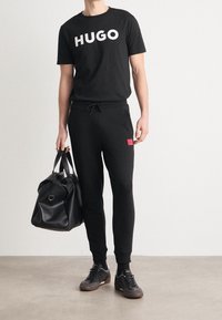 Hombre vistiendo un atuendo casual completamente negro con camiseta con el logo HUGO, pantalones jogger con parche de logo rojo, zapatillas negras y sosteniendo un bolso de viaje de cuero negro.