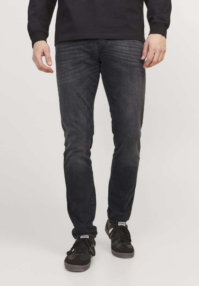 Jeans gris oscuro con un corte slim, que presentan un ligero desvanecimiento, tejido elástico y un diseño clásico de cinco bolsillos. Combinados con zapatillas deportivas negras.