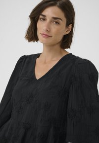 Schwarzes besticktes Kleid mit V-Ausschnitt, floralen Mustern, gerafften Ärmeln und einer fließenden Silhouette. Weicher, strukturierter Stoff.