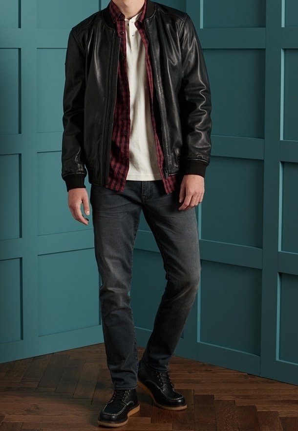 Homme portant une veste en cuir noire, une chemise à carreaux rouges, un t-shirt blanc, un jean foncé et des chaussures noires, debout contre un mur à panneaux bleu sarcelle.