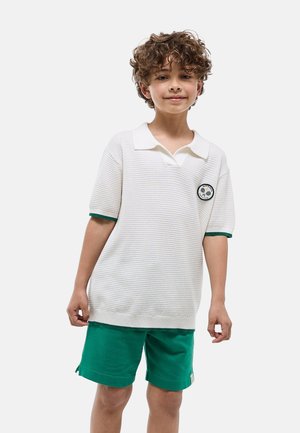 Garçon aux cheveux bouclés portant un polo tricoté blanc à manches courtes avec bordure verte et un short vert, debout devant un fond clair uni.