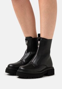 Botines negros hasta el tobillo con acabado en cuero liso, que presentan una cremallera diagonal y una suela gruesa para mayor agarre y estabilidad.
