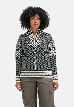 Kvinne med krøllete hår som har på seg en svart og hvit mønstret strikkegenser med halvlange glidelås og grå cargobukser på en ensfarget bakgrunn.