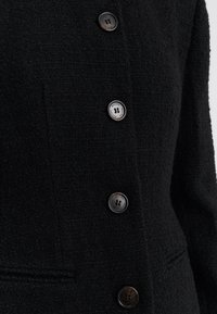 Fekete, texturált kabát, gombos záródással, négy kerek, sötét gombbal és oldalzseb részlettel. Az anyag puha, enyhén bouclé hatású.