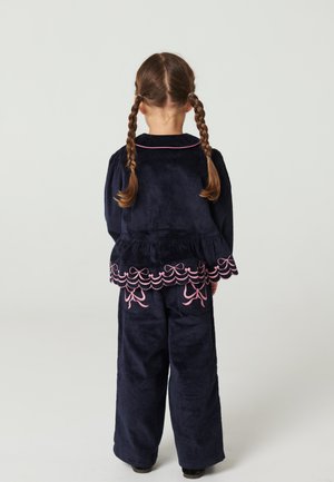 Enfant aux cheveux bruns tressés portant une tenue en velours côtelé bleu marine avec des nœuds brodés roses et des bords festonnés, debout de dos.