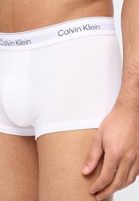 Κοντινό πλάνο ενός άνδρα που φοράει λευκά μποξεράκια Calvin Klein με ελαστική μέση με λογότυπο, το χέρι του ακουμπάει στο μηρό του.