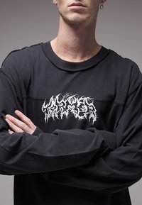 Μαύρο μακρυμάνικο πουκάμισο με στρογγυλή λαιμόκοψη, που διαθέτει ένα γραφικό λογότυπο σε λευκό. Το ύφασμα φαίνεται μαλακό με εμφανείς ραφές και χαλαρή εφαρμογή.