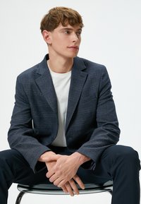 Marinblå blazer med en subtil rutmönster, tillverkad av textil med struktur, visad på en sittande modell, kombinerad med en vit t-shirt och marinblå byxor.