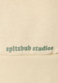 Hellbeige Stoff mit einer gestrickten Textur, auf dem nahe der unteren Kante ein auffälliges grünes Textlogo mit der Aufschrift "spitzbub studios" erscheint.