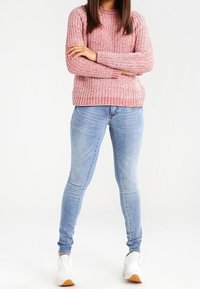Suéter de punto rosa con cuello redondo, combinado con jeans skinny azul claro y zapatillas blancas con acentos en marrón, de pie sobre un fondo liso.
