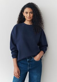 Teget plava majica s okruglim izrezom i opuštenim krojem, s rebrastim manžetama. Uparena s trapericama srednjeg pranja.