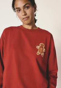 Rød sweatshirt med lange ærmer og rund hals, med et pebersvends patch i brunt med hvid og rød detaljering på fronten.