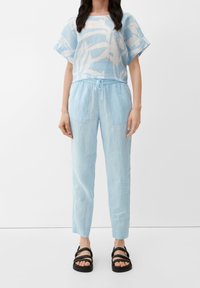 Lichtblauw linnen shirt met abstract wit ontwerp, gecombineerd met bijpassende lichtblauwe linnen broek met een elastische tailleband en zijzakken. Zwarte sandalen.