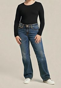 Personne portant un haut noir à manches longues, un jean droit bleu et des baskets blanches, debout devant un fond beige uni.