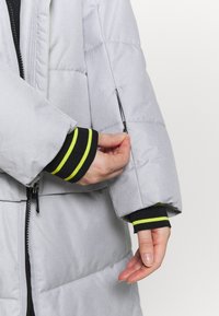 Main avec poignet rayé noir et jaune fermant une petite poche zippée sur la manche d'une veste matelassée gris clair.