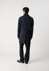 Casaco tailorizado azul-marinho com gola alta, mangas longas e um design de costas retas. Usado sobre calças pretas e sapatos.