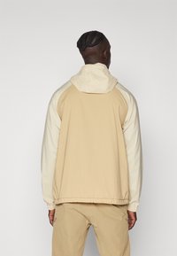 Chaqueta con capucha en color beige y crema, con cordón ajustable, puños elásticos y un corte relajado. Tejido suave con un acabado mate.