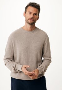 Beige, strukturierter Strickpullover mit Rundhalsausschnitt und langen Ärmeln. Verfügt über gerippte Bündchen und eine lockere Passform, getragen über dunklen Hosen.
