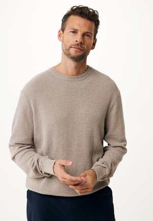 Jersey de punto texturizado beige con cuello redondo y mangas largas. Presenta puños acanalados y un ajuste relajado, llevado sobre pantalones oscuros.