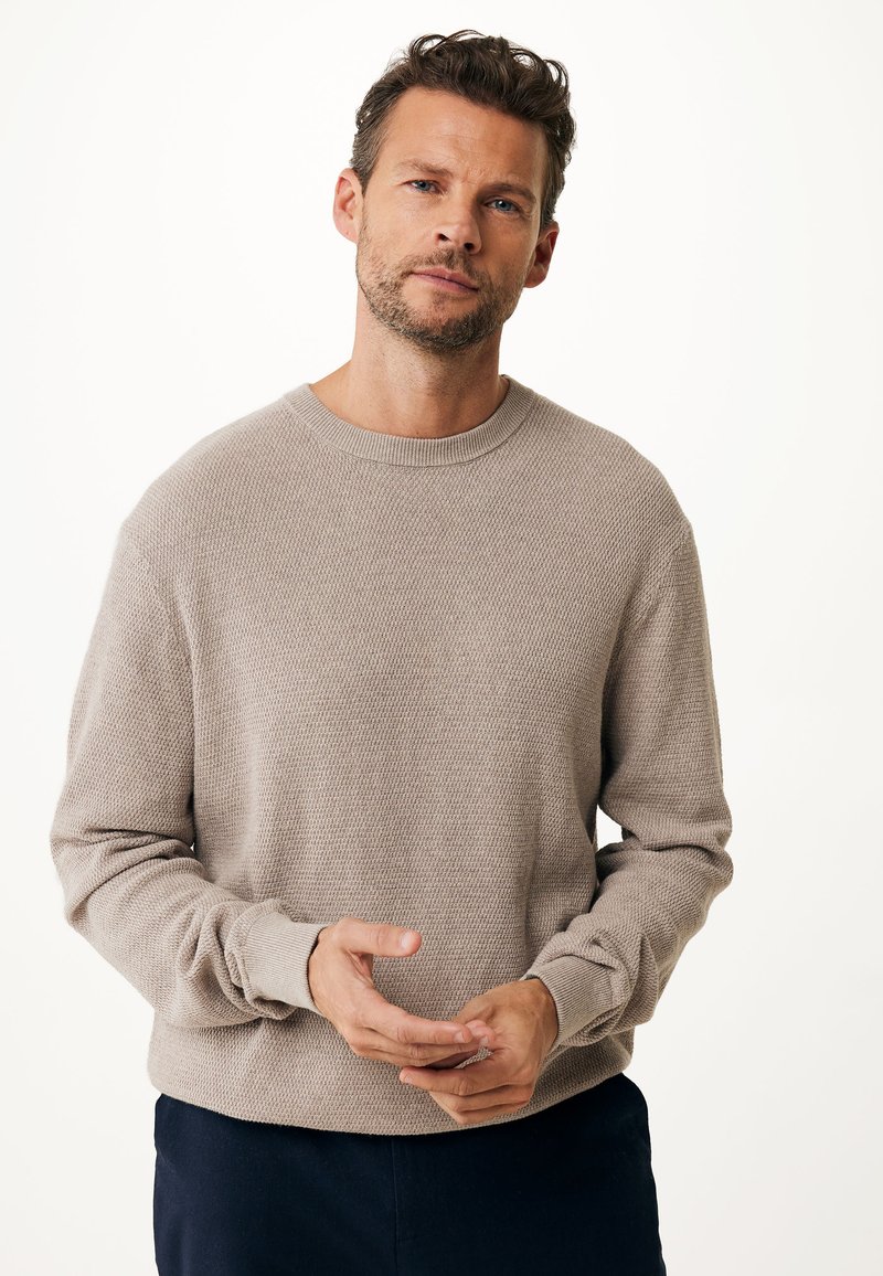 Beige, strukturierter Strickpullover mit Rundhalsausschnitt und langen Ärmeln. Verfügt über gerippte Bündchen und eine lockere Passform, getragen über dunklen Hosen.