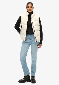 Smanicato trapuntato color avorio, con collo alto, chiusura con zip e due tasche laterali, abbinato a un maglione nero lavorato a maglia e jeans blu.