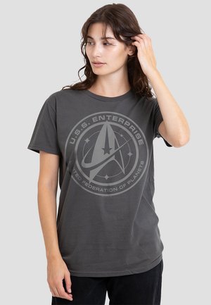 Woman with brown hair adjusting it, wearing a gray U.S.S. Enterprise t-shirt with a circular space-themed emblem.