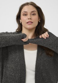 Siv cardigan z ribami z dolgimi rokavi, ki ima luknje za palec na manšetah, oblečen čez belo prilegajočo majico. Mehka, teksturirana tkanina.