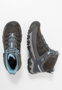 Buty wykonane z ciemno-brązowego materiału syntetycznego z szarymi akcentami, z teksturowaną podeszwą i jasnoniebieskimi sznurowadłami. Wysoki krój zapewniający wsparcie.