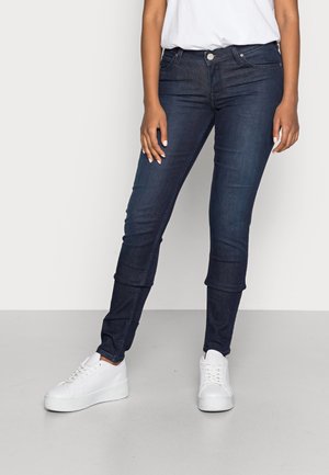 Dark blue skinny jeans in stretch denim, featuring a mid-rise waist, front pockets, and a cuffed hem, paired with white sneakers.