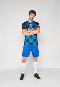 Black and blue checkered soccer jersey with short sleeves, paired with solid blue shorts and white socks, worn with black soccer cleats.