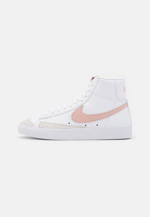 Hvid højtop sneaker med lyserød Nike swoosh og snørebånd, med tekstureret tåhætte og flad hvid sål, sidevisning på ensfarvet baggrund.
