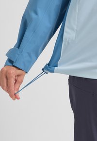 Giacca leggera blu e azzurra con cordino regolabile all'orlo, maniche lunghe e dettagli con polsini abbottonati. Tessuto liscio.