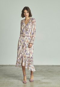 Robe fleuri avec des volants, manches longues et un ourlet fluide. Le tissu présente un fond crème clair avec des fleurs violettes et roses.