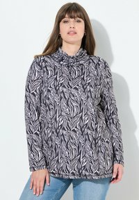 Haut à manches longues avec un col drapé, en tissu noir orné de motifs de feuilles blanches. Accompagné de jeans en denim bleu clair.