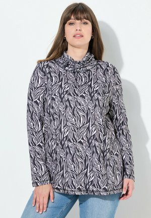 Haut à manches longues avec un col drapé, en tissu noir orné de motifs de feuilles blanches. Accompagné de jeans en denim bleu clair.