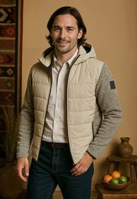 Beige gewatteerde jas met een grijze gestructureerde mouw en capuchon, voorzien van een zwart accentpatch. Gedragen over een wit shirt en een donkere spijkerbroek.