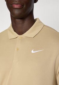 Nehéz, sima anyagból készült bézs polo ing, klasszikus gallérral, két gombos nyakkal, és a bal oldalon található fehér Nike log�óval.