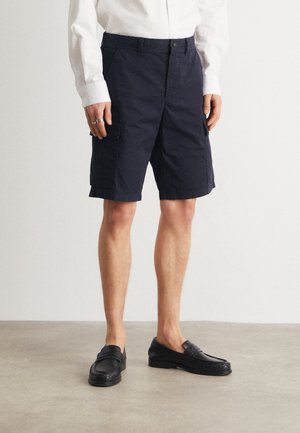 Uomo che indossa pantaloni cargo blu navy, camicia a maniche lunghe bianca, mocassini neri, in piedi su un pavimento chiaro e liscio di fronte a un muro semplice.