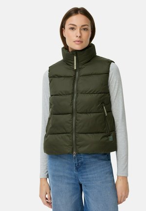 Junge Frau in einer dunkelgrünen, gesteppte ärmellosen Weste über einem hellgrauen Langarmshirt und blauen Jeans, die vor einem schlichten Hintergrund steht.