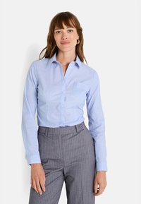 Camisa de color azul claro ajustada con un cuello clásico, mangas largas, botones frontales y un bolsillo en el pecho izquierdo, combinada con pantalones de rayas grises.