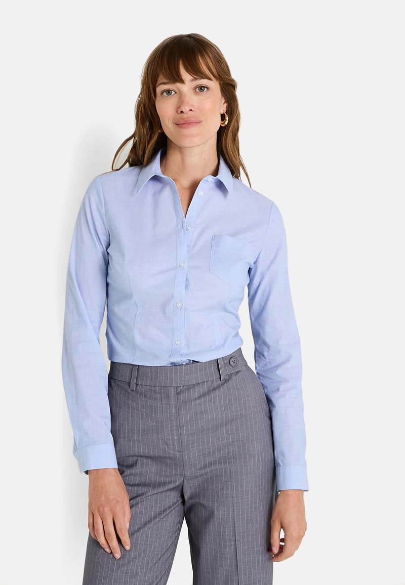 Camisa de color azul claro ajustada con un cuello clásico, mangas largas, botones frontales y un bolsillo en el pecho izquierdo, combinada con pantalones de rayas grises.